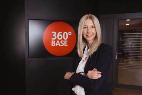 Lächelnder Mitarbeiter im Anzug mit 360°-BASE-Pin; im Hintergrund spielen Kollegen Tischtennis.