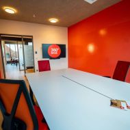 Arbeitsraum mit Besprechungstisch, roten Stühlen, Monitor und Blick ins Nachbarbüro