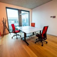 Büroraum mit großem Tisch, drei roten Stühlen, Fensterfront mit Tageslicht und roter Wand