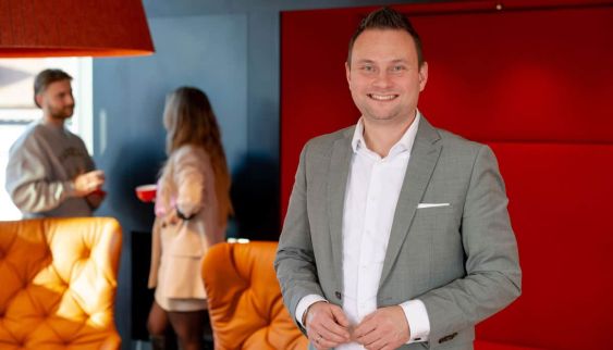 Lächelnder Berater in Lounge mit orangefarbenen Sesseln; im Hintergrund locker plaudernde Kolleg:innen, offene Atmosphäre.